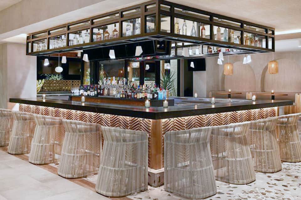 Elegant indoor bar with black counter, woven bar stools, decorative overhead shelving, and modern pendant lighting
