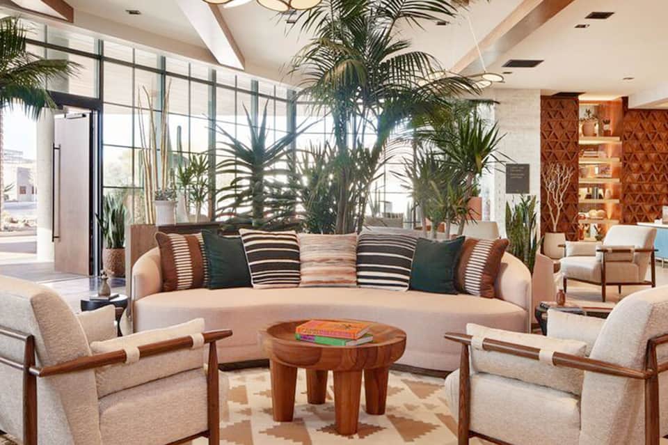 Contemporary hotel lobby with curved cream sofa, striped pillows, potted palm plants, and large windows.