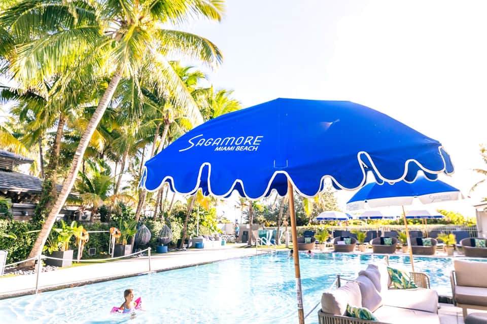 Pool area with blue Sycamore Miami Beach umbrella, palm trees, and guests enjoying water activities