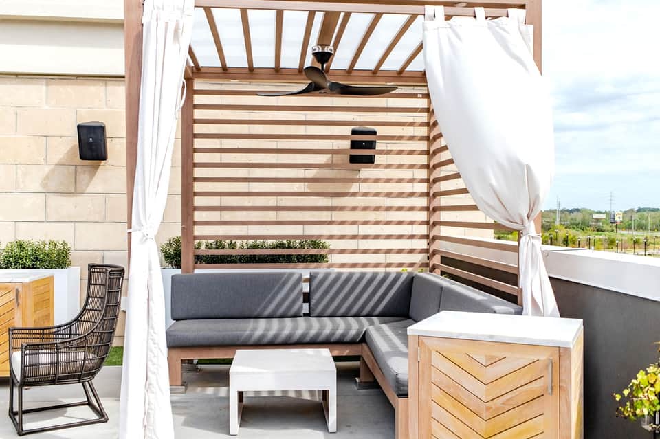 Modern outdoor living space with gray sectional sofa under wooden pergola with white curtains and ceiling fan