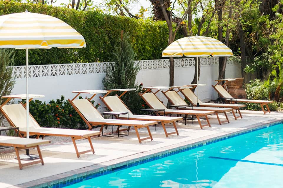 Resort pool deck with yellow and white striped umbrellas, wooden lounge chairs, and white privacy fence