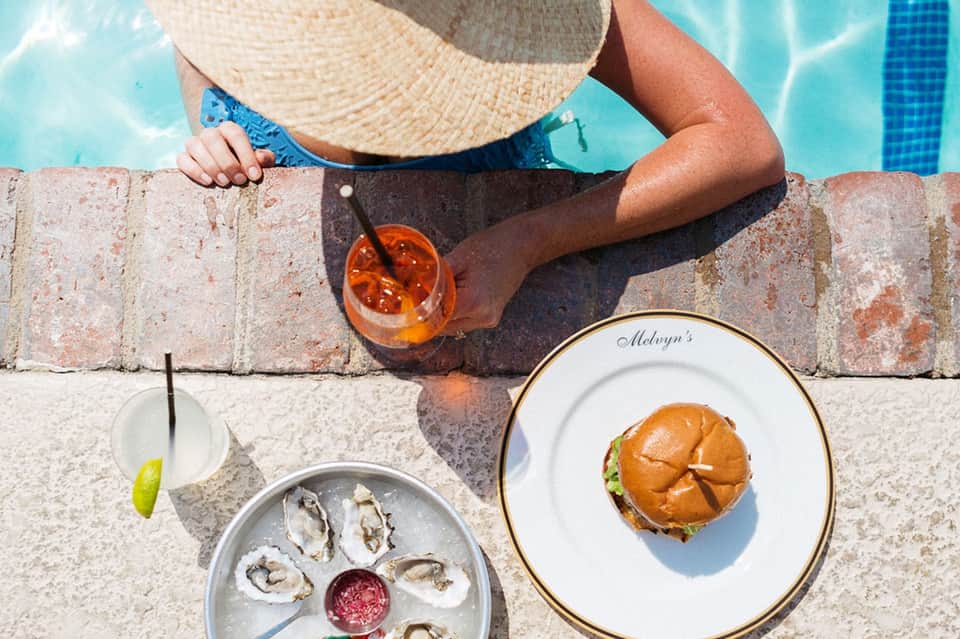 Person in sun hat poolside holding tropical cocktail with oysters and burger plates nearby