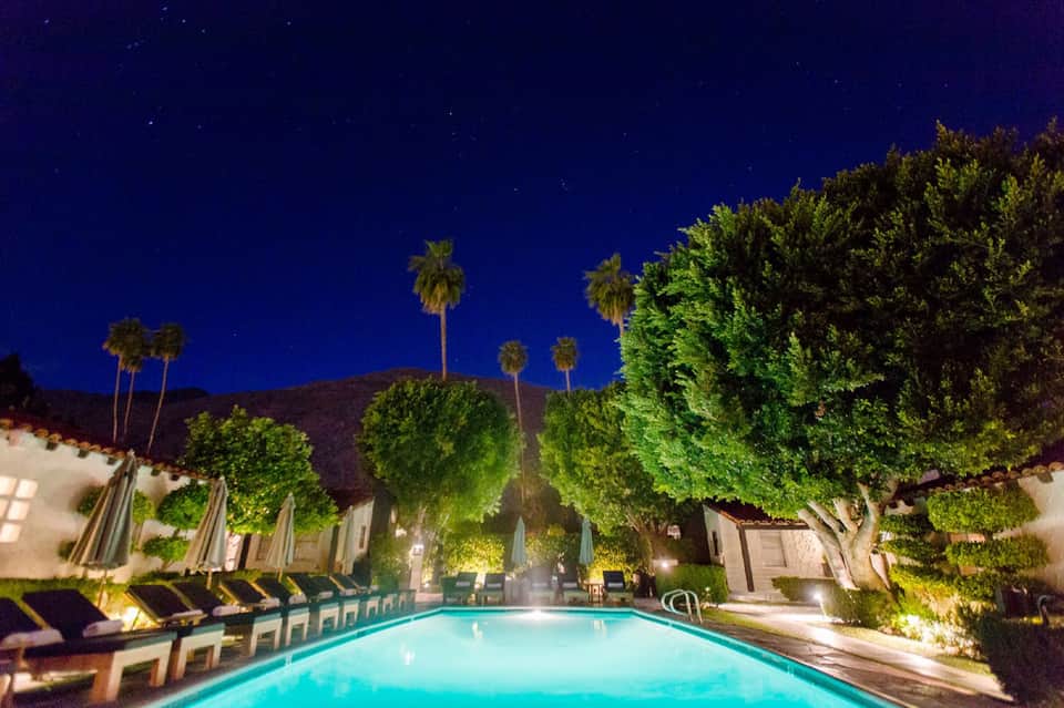 Illuminated resort pool at night with palm trees, loungers, and starry sky overhead