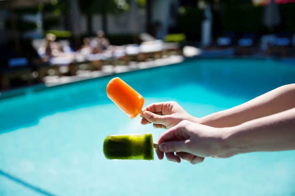 Hands holding colorful popsicles over a bright turquoise swimming pool