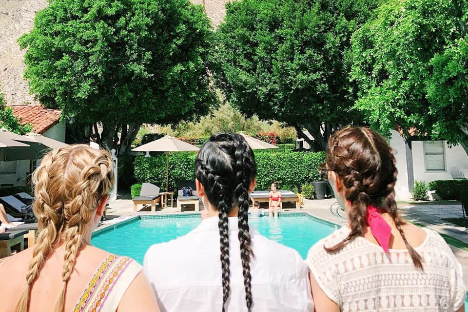 Three girls with braided hairstyles viewing outdoor swimming pool surrounded by mature trees and umbrellas