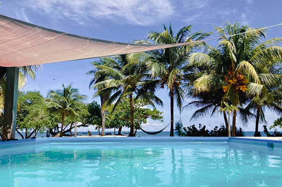 Resort pool with sail shade canopy, palm trees, hammock, and tropical ocean view under blue sky