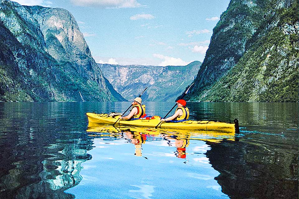 Two kayakers paddling yellow kayak through calm fjord surrounded by dramatic cliff mountains