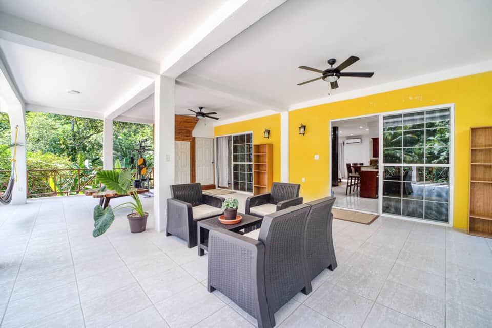 Covered patio with yellow accent wall, wicker seating, ceiling fans, and tropical landscaping