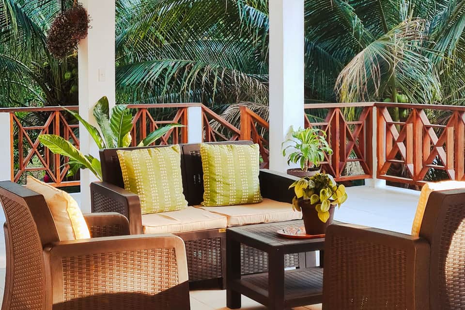 Tropical terrace lounge with wicker furniture, yellow pillows, palm trees, and wooden railings