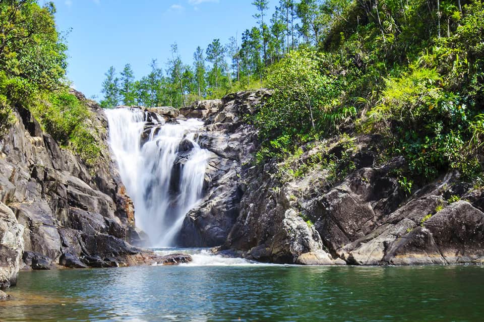 Scenic waterfall cascading over dark rocks into turquoise pool surrounded by lush green forest and vegetation