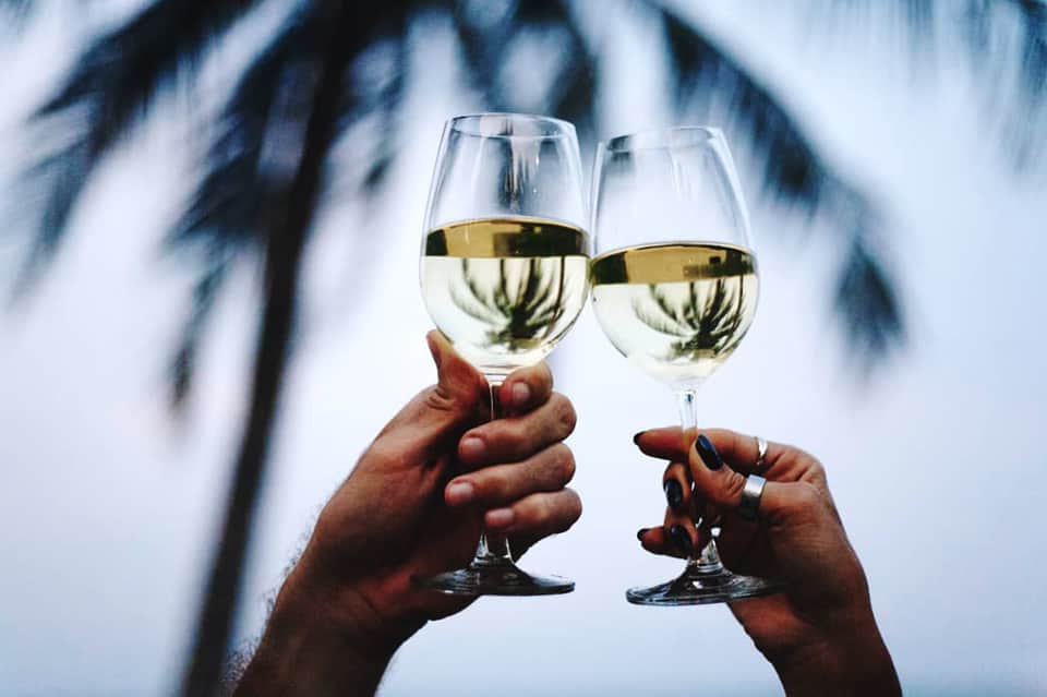 Two people toasting with white wine glasses against a tropical backdrop
