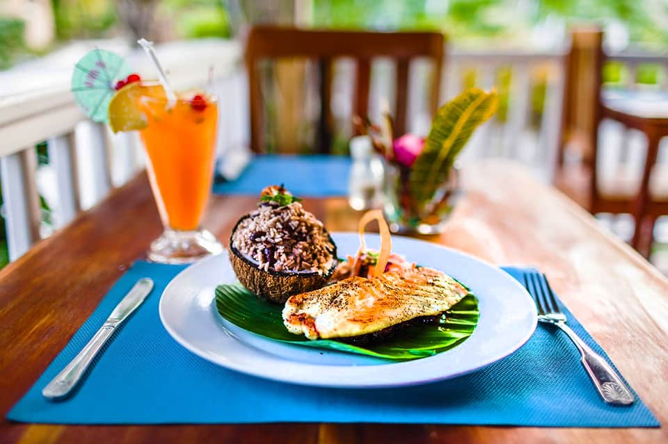 Beachfront dining table with grilled fish, stuffed coconut, tropical orange cocktail, and water view