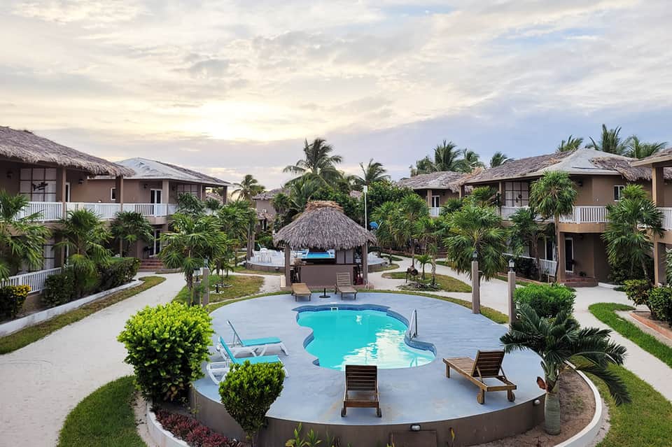Resort compound featuring thatched-roof bungalows, central swimming pool with lounge chairs, and manicured gardens