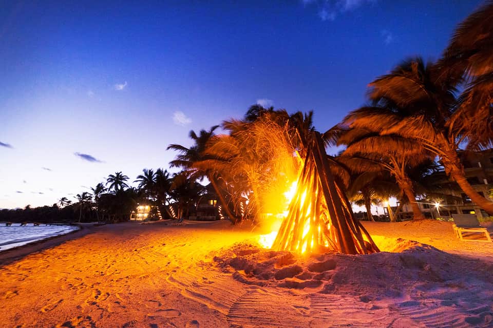 Bonfire on beach at dusk with palm trees and lit buildings in background