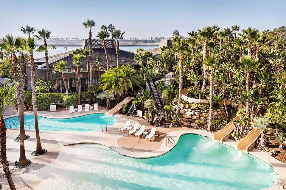 Tropical resort pool complex with turquoise water, palm trees, waterslide, and river view in background
