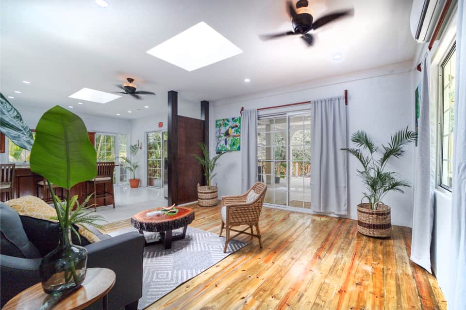 Bright living room with tropical plants, wooden floors, ceiling fans, and glass doors opening to outdoor patio