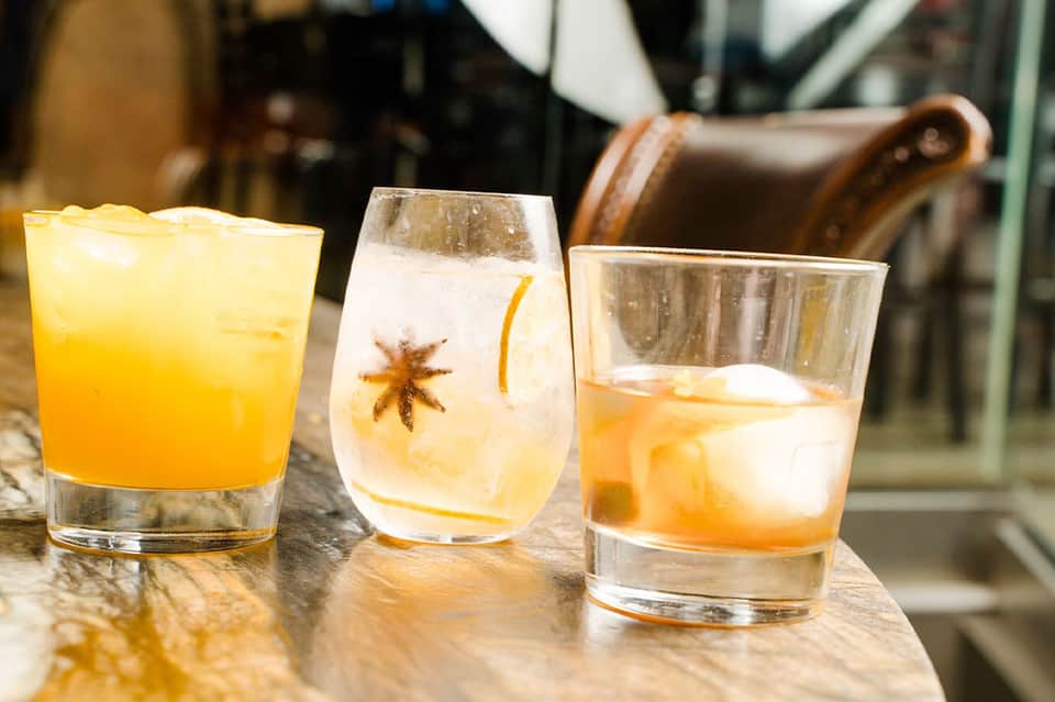 Three citrus-colored cocktails on marble bar counter with leather chair in background