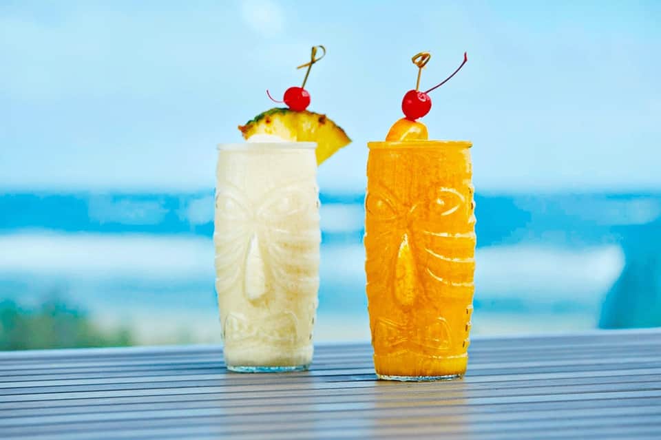 Two tropical cocktails with pineapple and cherry garnish overlooking ocean and beach