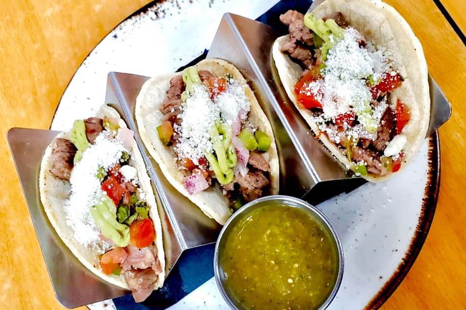 Three tacos with meat, fresh vegetables, cheese, and salsa verde on white plate