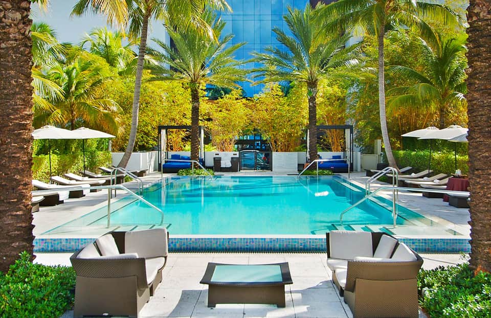 Luxury resort pool with turquoise water, palm trees, white umbrellas, and lounge chairs surrounded by modern architecture
