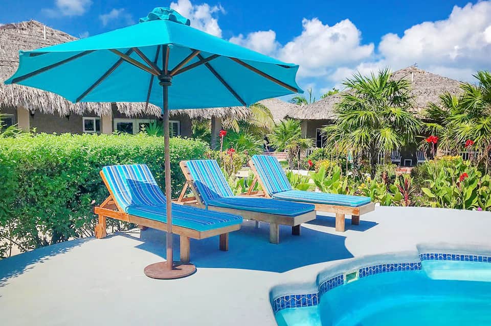 Tropical resort poolside with turquoise umbrella, wooden loungers, palm trees, and thatched-roof buildings
