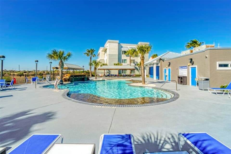 Resort pool area with circular design, palm trees, lounge chairs, and modern white building