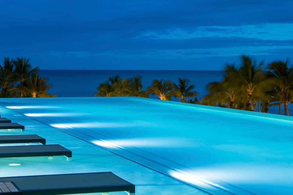 Illuminated infinity pool overlooking ocean at dusk with palm trees and lounge chairs