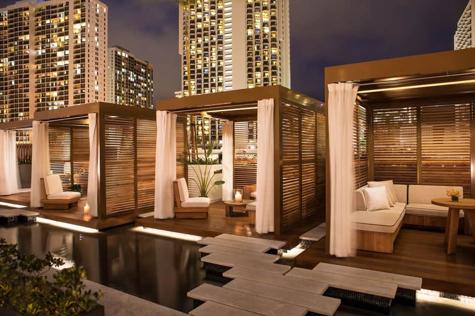 Rooftop lounge area at night with wooden pergolas, white curtains, seating areas, and city skyline backdrop