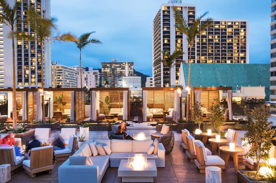 Upscale rooftop lounge at dusk with fire pits, modern seating, and illuminated high-rise hotels