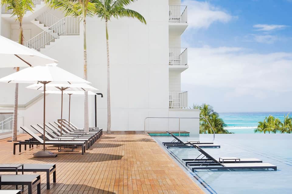Resort pool deck with white umbrellas, lounge chairs, and modern building overlooking tropical ocean