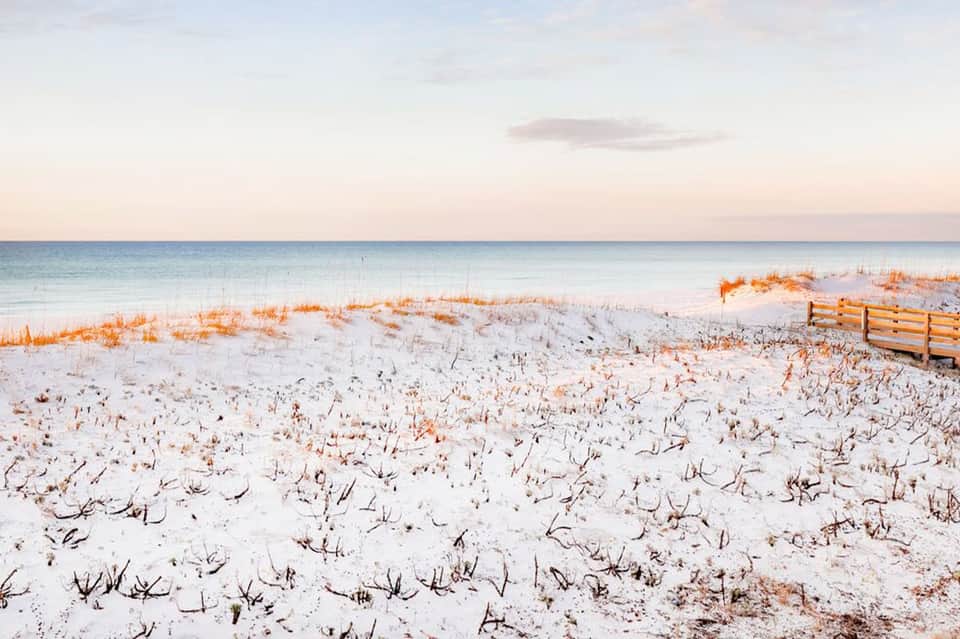 Serene beach at sunset with white sand, sea grass, wooden fence, and calm ocean waters