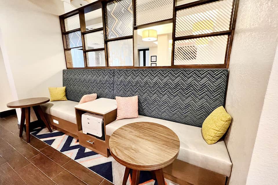 Modern hotel lobby lounge with geometric patterned headboard, wooden tables, and pastel accent pillows.
