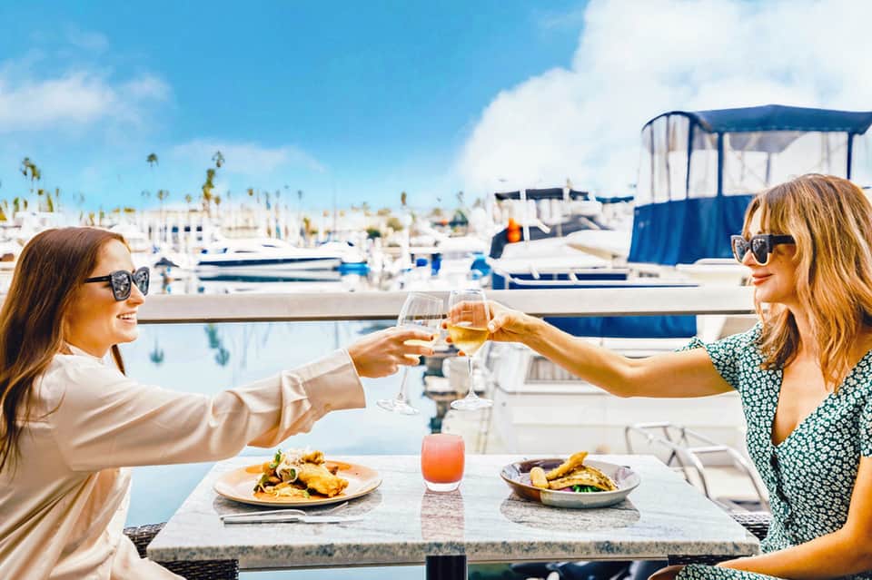 Two women toasting wine glasses at waterfront restaurant overlooking marina