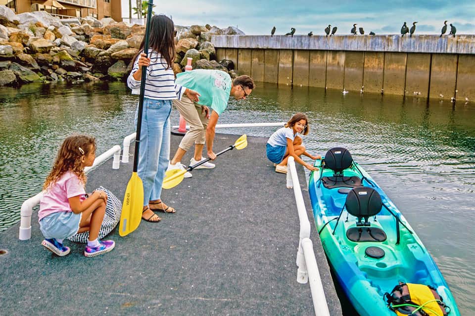 Family of four preparing to kayak, holding paddles on dock with turquoise water and birds in background