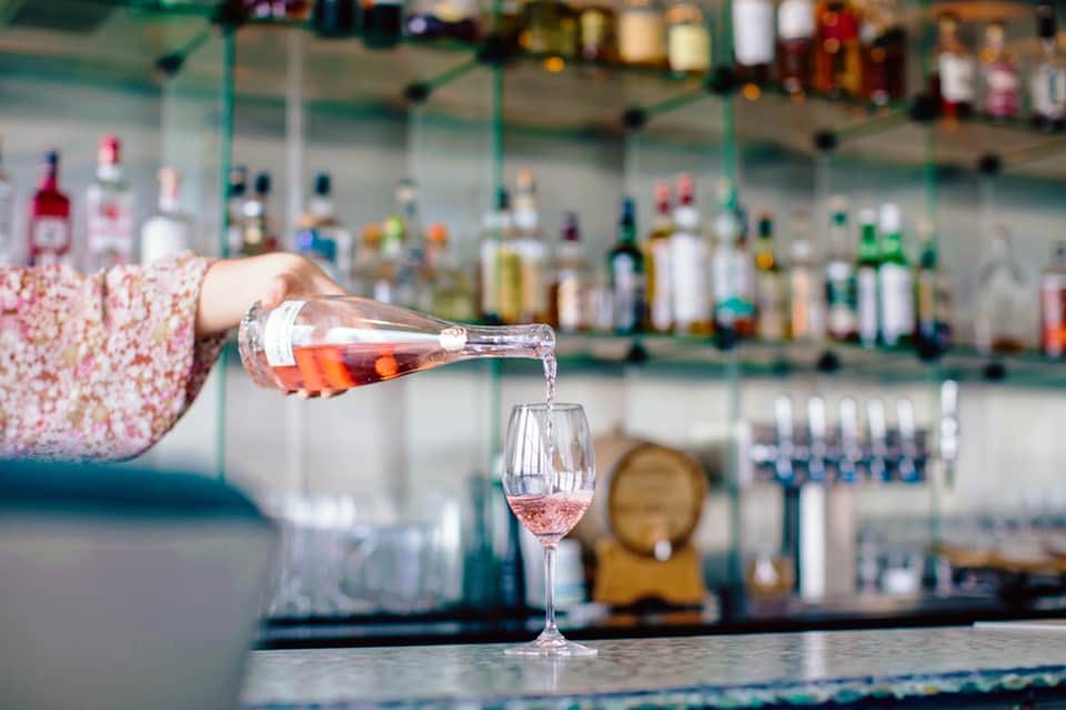 Bartender pouring rosé wine into glass at bar with bottles in background