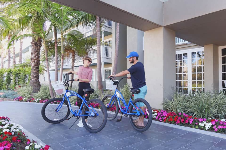 Two people riding blue bicycles near resort building with palm trees and colorful flower beds