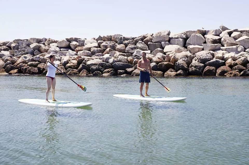 Two people paddleboarding on calm water near rocky breakwater