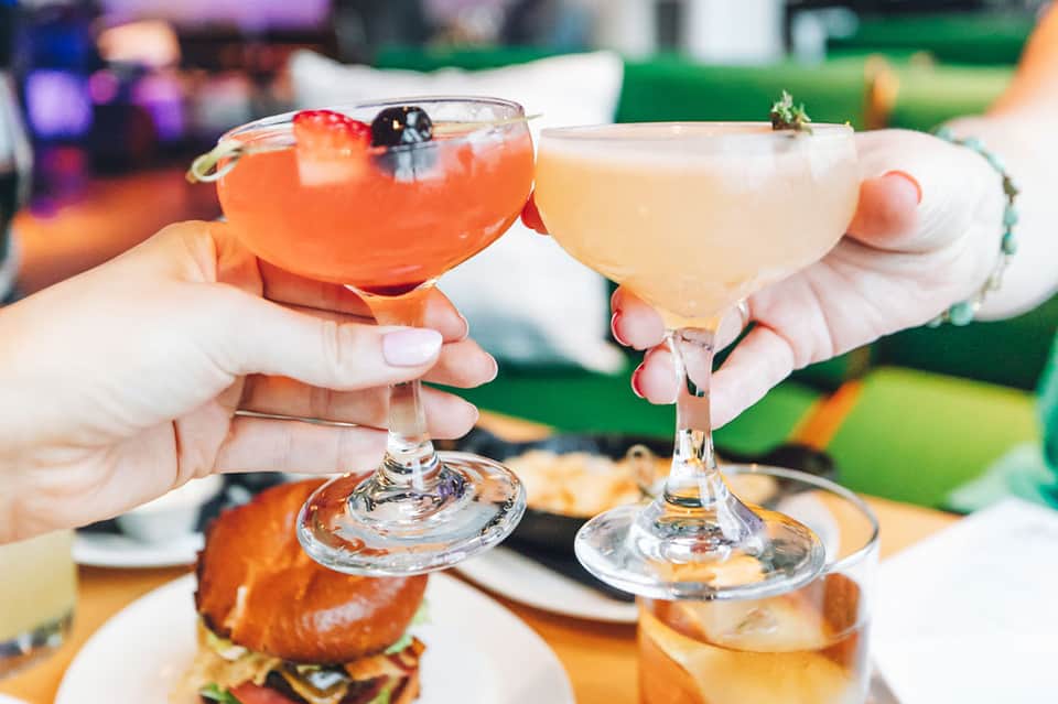 Two hands toasting with colorful cocktails at an outdoor event with food and decorations