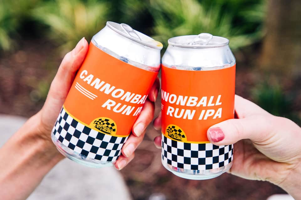 Two hands holding Cannonball Run IPA orange cans with checkered pattern design outdoors