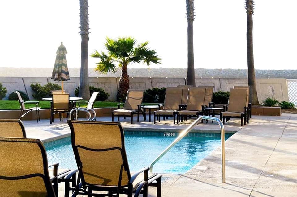 Poolside lounge area with tan chairs, umbrellas, palm trees, and mountain views in background