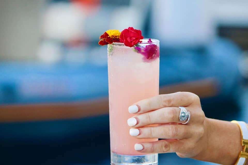 Pink cocktail with edible flowers garnish held by hand with ring, ocean backdrop