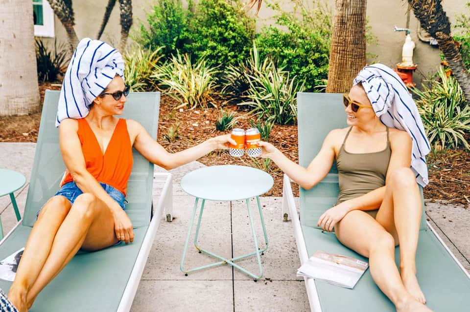 Two women in colorful beach attire toasting with drinks in courtyard lounge chairs