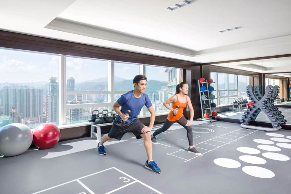 Two people exercising in a high-rise gym with floor-to-ceiling windows overlooking a cityscape and mountains