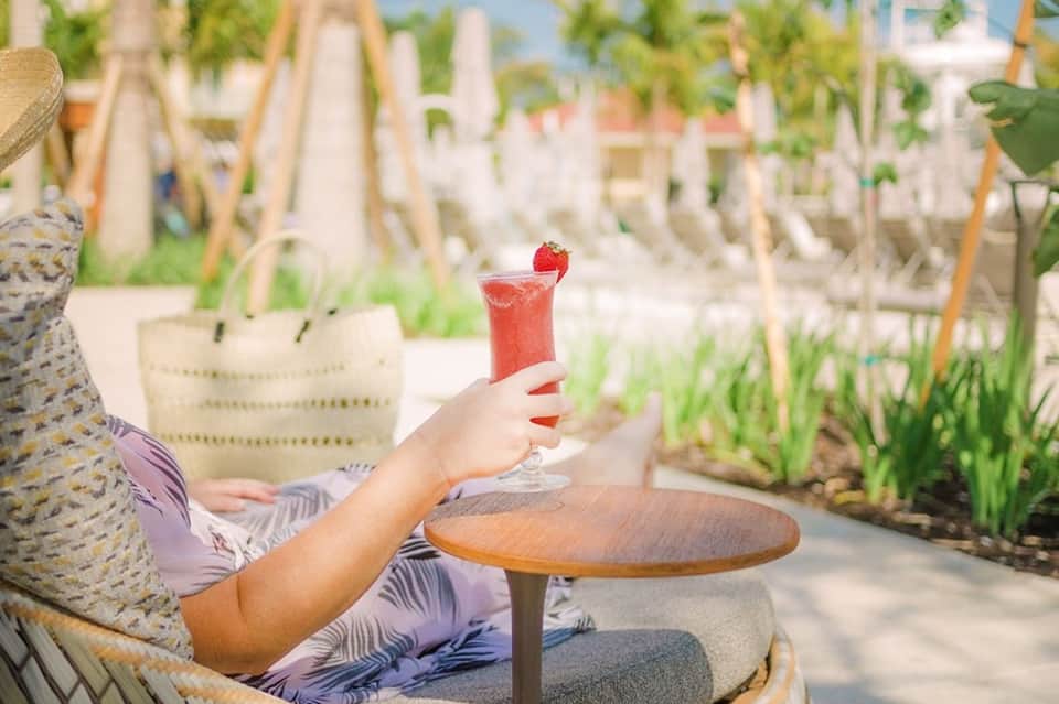 Guest holding strawberry cocktail overlooking beach cabanas and lounge chairs on sandy resort grounds