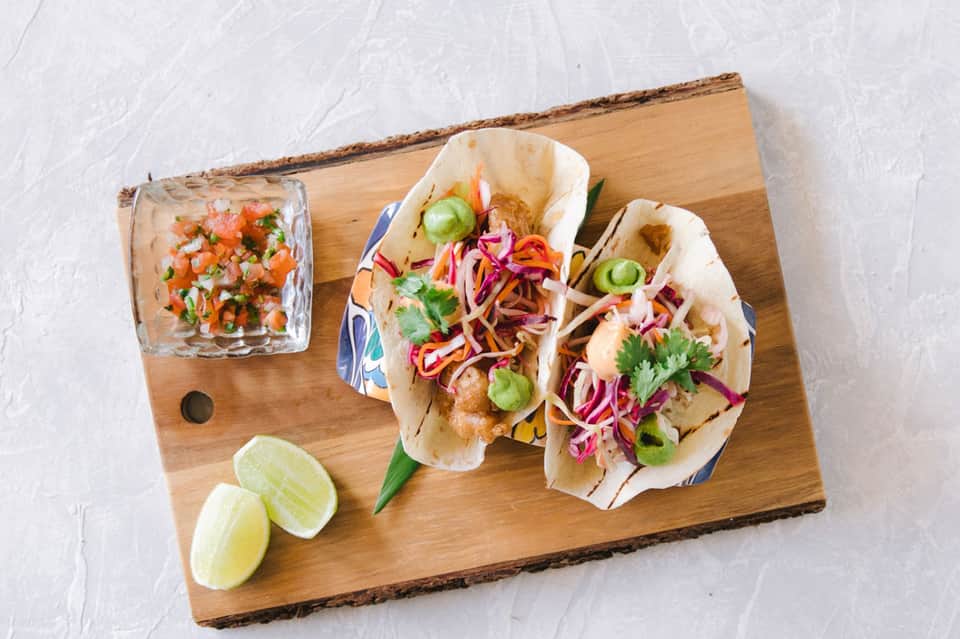 Gourmet tacos on wooden board with cilantro, pickled onions, lime wedges, and salsa