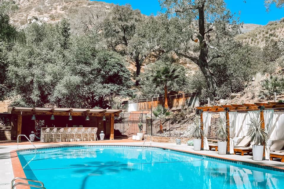 Resort pool surrounded by lounge chairs, wooden pergolas, and olive trees on hillside