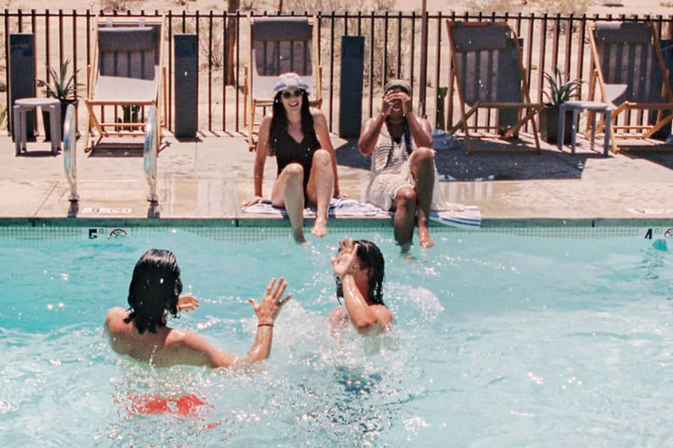 Resort pool with guests swimming, spectators on wooden deck chairs, and beach cabanas in background