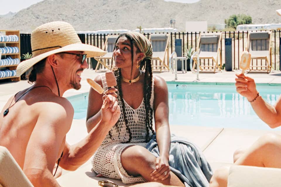 Man and woman enjoying drinks poolside at desert resort with cabanas and mountains