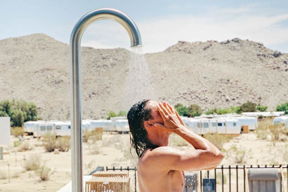 Shirtless man rinsing off under an outdoor shower in a desert setting with mountains and trailers