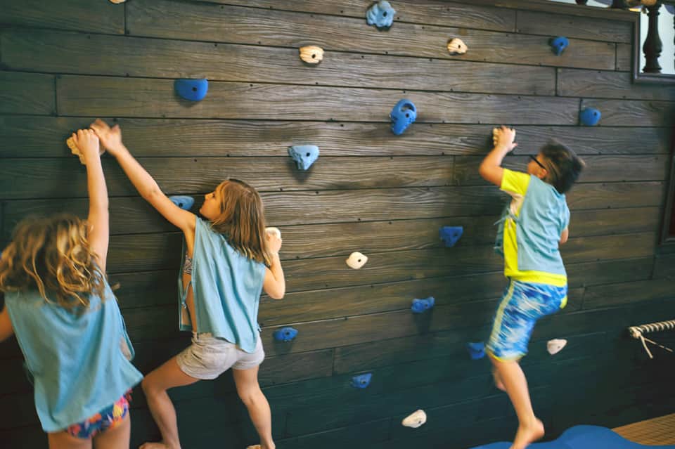 Children climbing a wooden wall with blue and white hand holds at an indoor activity center